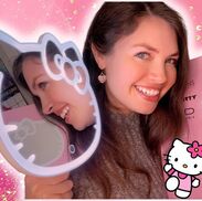 Lesley is smiling with heart-shaped dangling earrings. She is holding the Hello Kitty makeup mirror in one hand and the box it came in on the other.